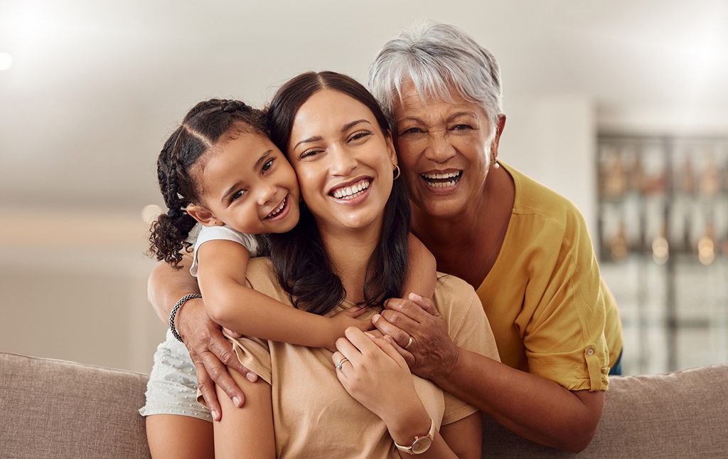 Grandmother, mom and child hug in a portrait for mothers day on a house sofa as a happy family in Colombia. Smile, mama and elderly woman love hugging young girl or kid and enjoying quality time